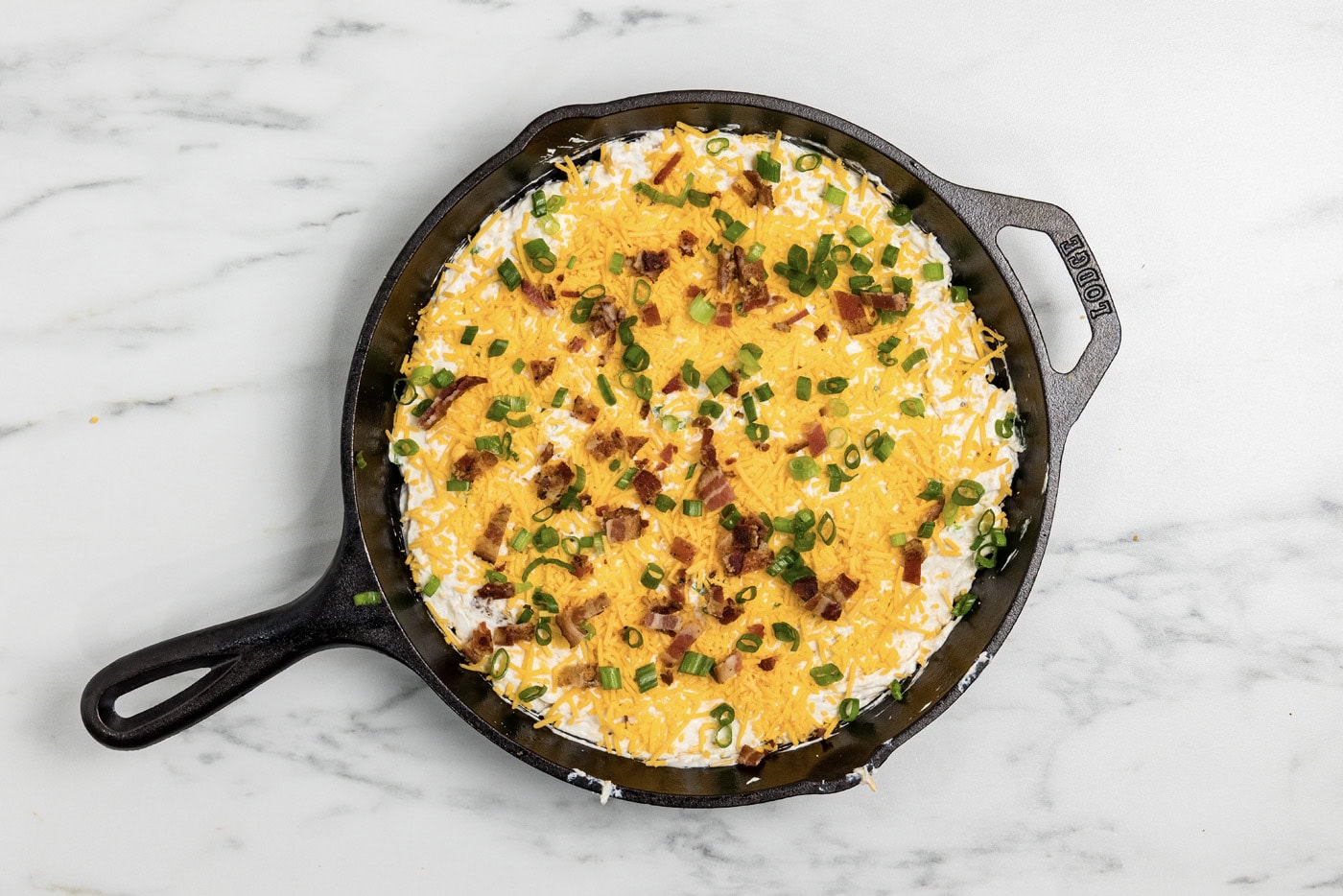 Dip covered with shredded cheddar, crumbled bacon, and sliced green onions in a cast iron skillet