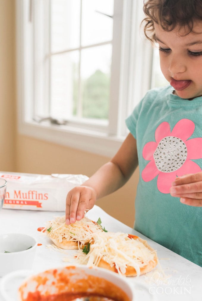 small child adding cheese to english muffin pizza