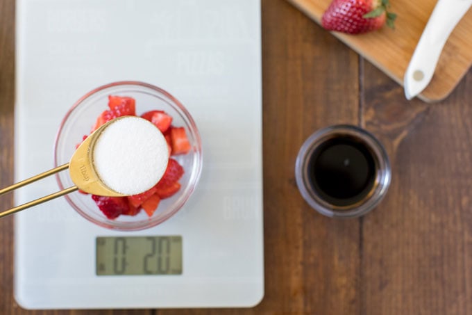 measuring spoon with sugar hovering over bowl of strawberries on a kitchen scale
