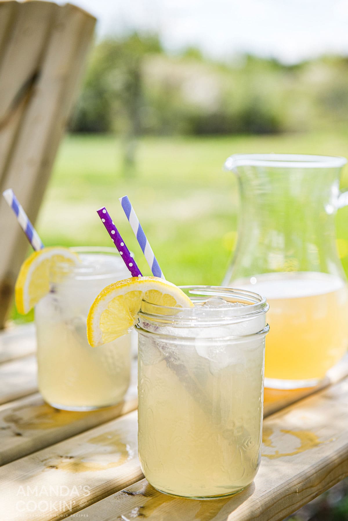 A pitcher of Lynchburg lemonade and two mason jars resting on a picnic table. Mason jars have a wedge of lemon and a straw in each glass.