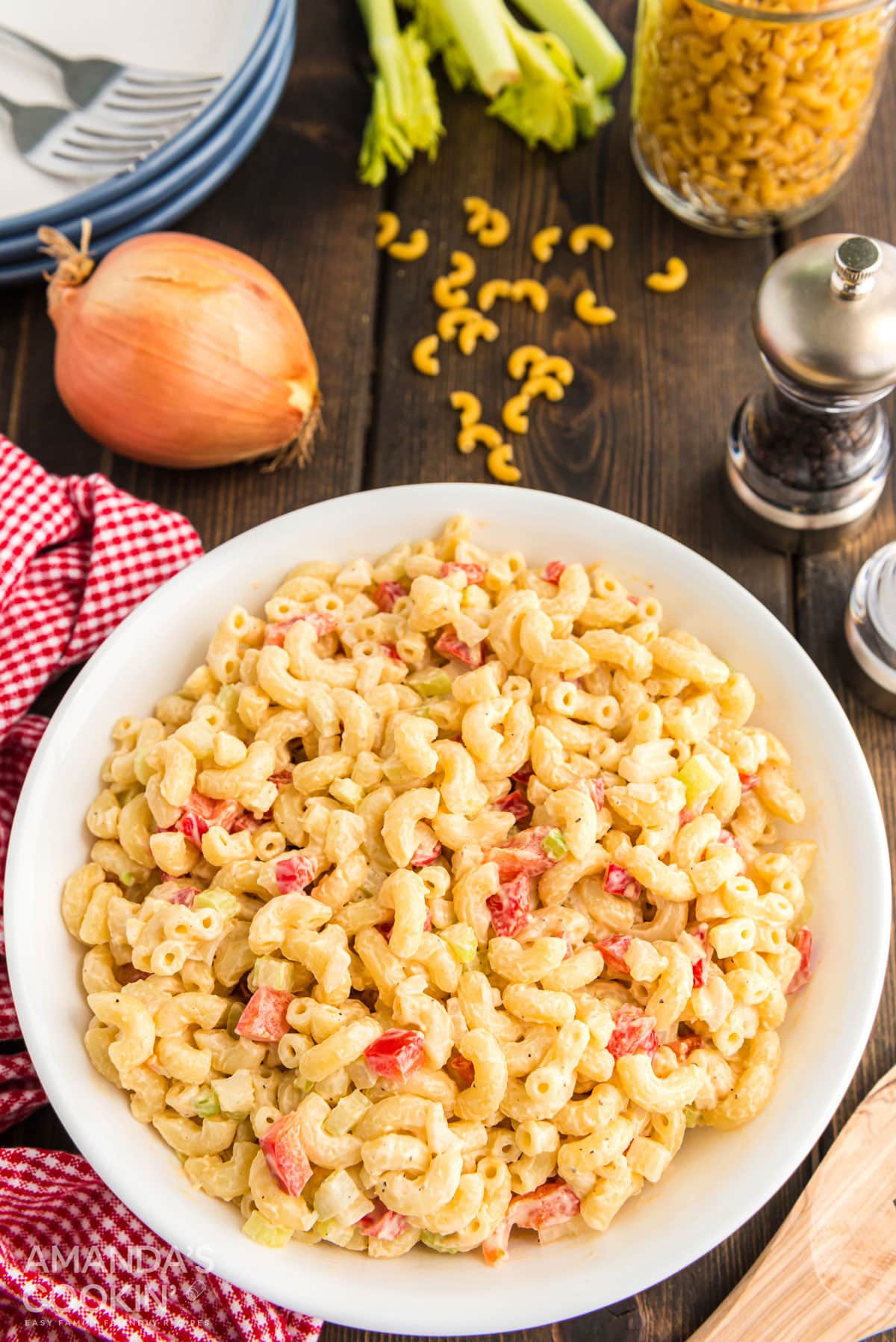 bowl of macaroni salad on a table with pepper, onion, dried pasta, and plates in background
