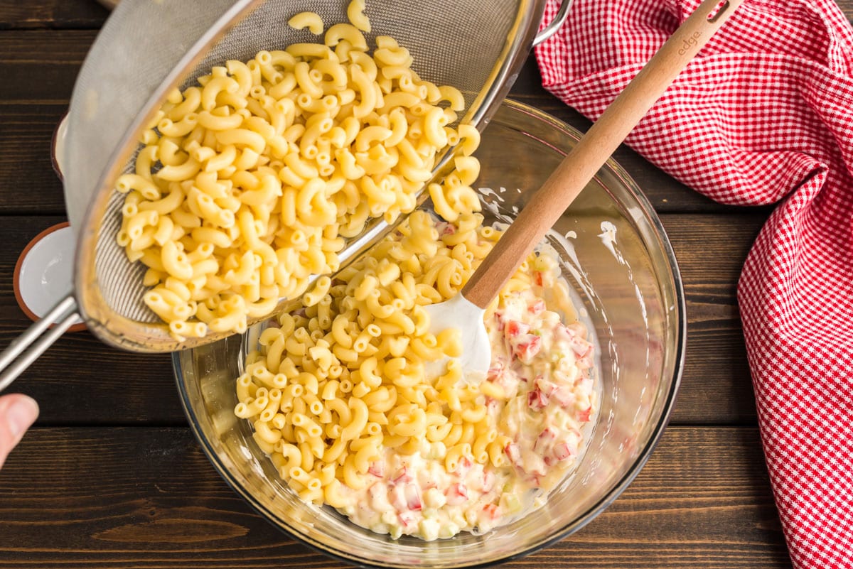 adding cooked macaroni to the pasta dressing and veggies in bowl