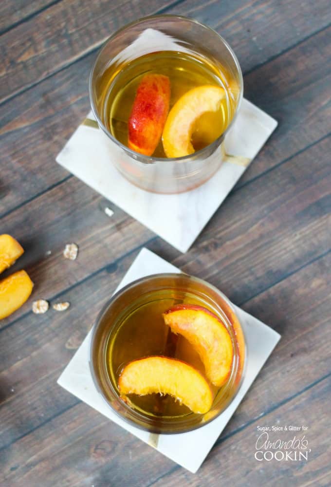 An overhead of two short clear glasses filled with peach pie cocktail with two peach slices floating on top.