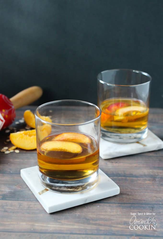 Two short clear glasses filled with peach pie cocktail with two peach slices floating on top.