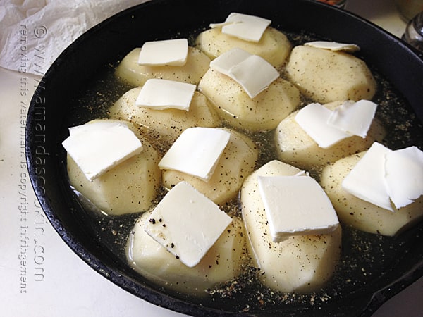 A close up photo of peeled potatoes in a cast iron skillet with slices of butter resting on top.