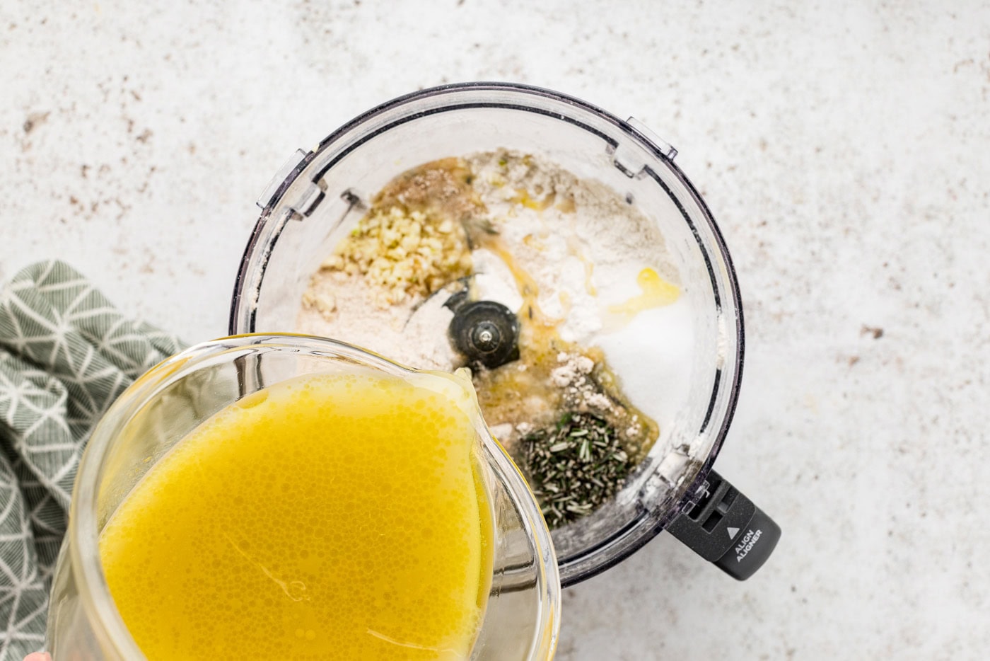 Pouring water, oil, and yeast mixture into food processor with flours, garlic, and rosemary