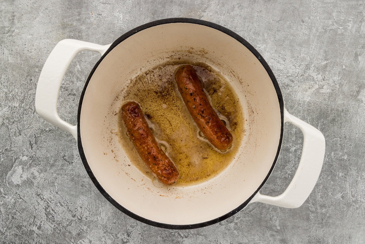 Two chicken sausage links browning in oil and butter in a white Dutch oven