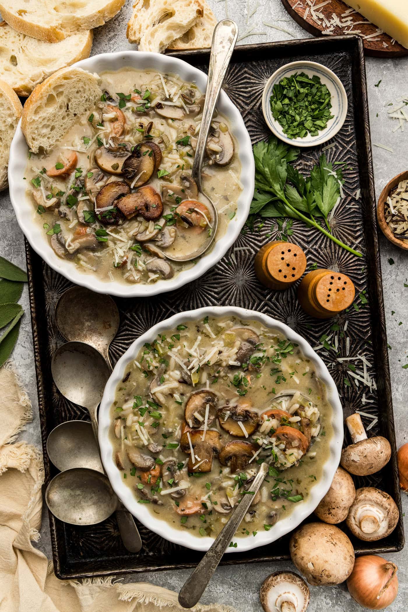 Two bowls of sausage mushroom wild rice soup on a dark metal tray with crusty bread, chopped parsley and whole mushrooms