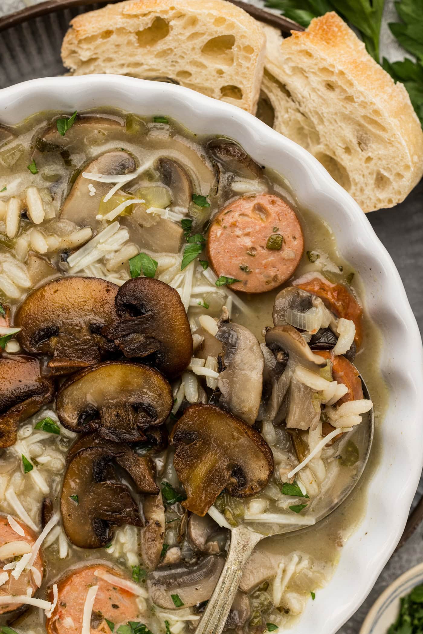 Close up of a bowl of sausage wild rice soup topped with browned mushrooms, grated Parmesan and parsley beside crusty bread