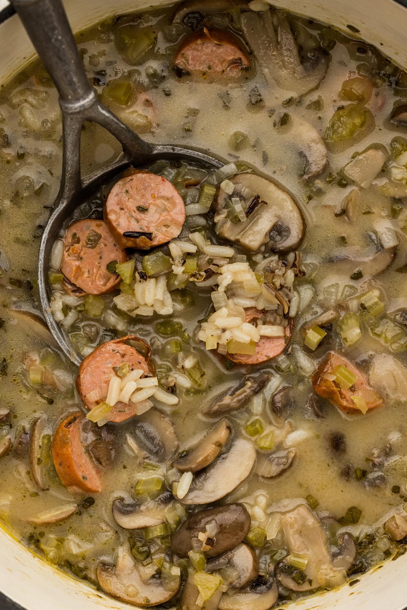 A ladle lifting sliced sausage, mushrooms and wild rice out of a pot of soup