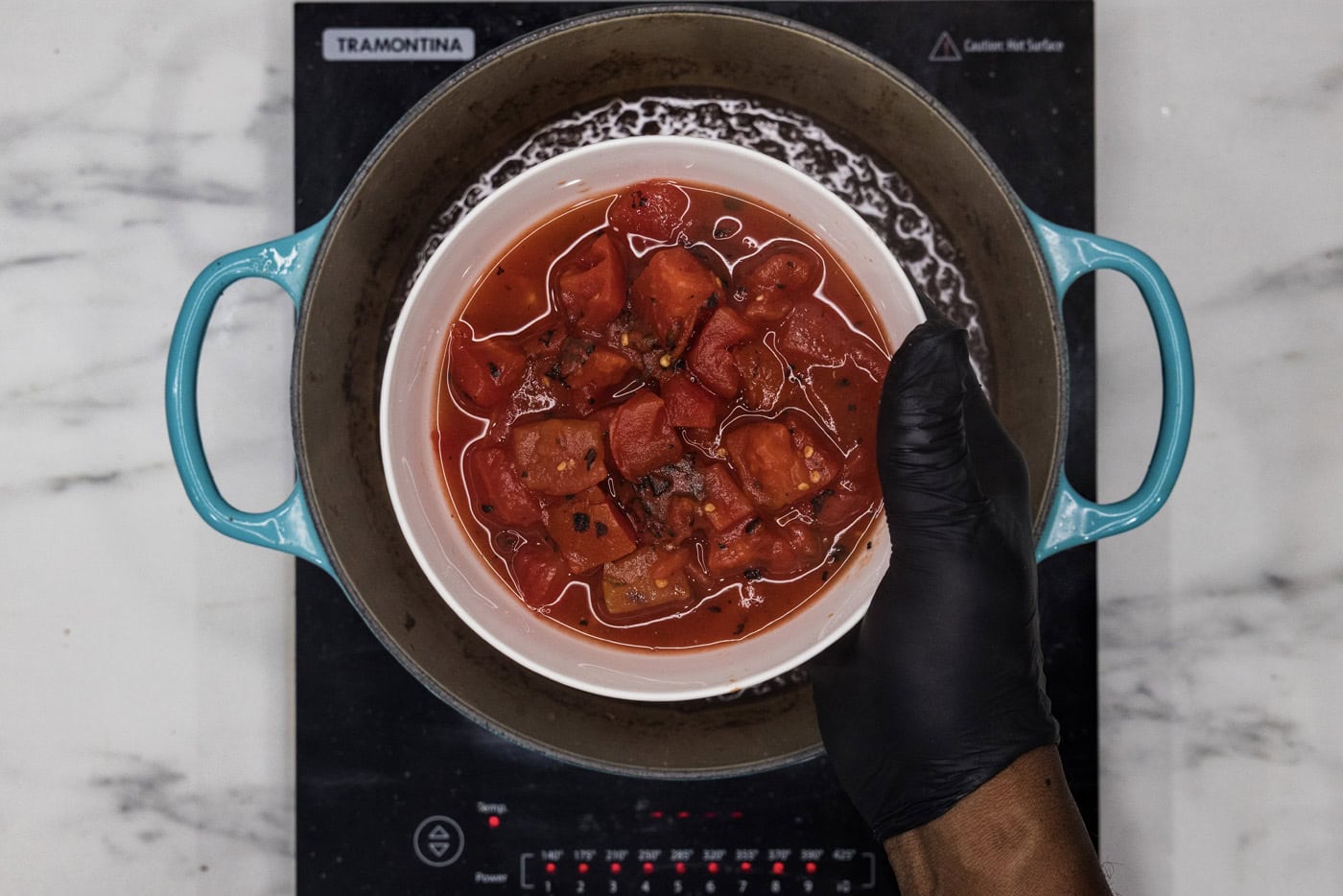 hand holding diced tomatoes in a bowl over dutch oven with braising liquid