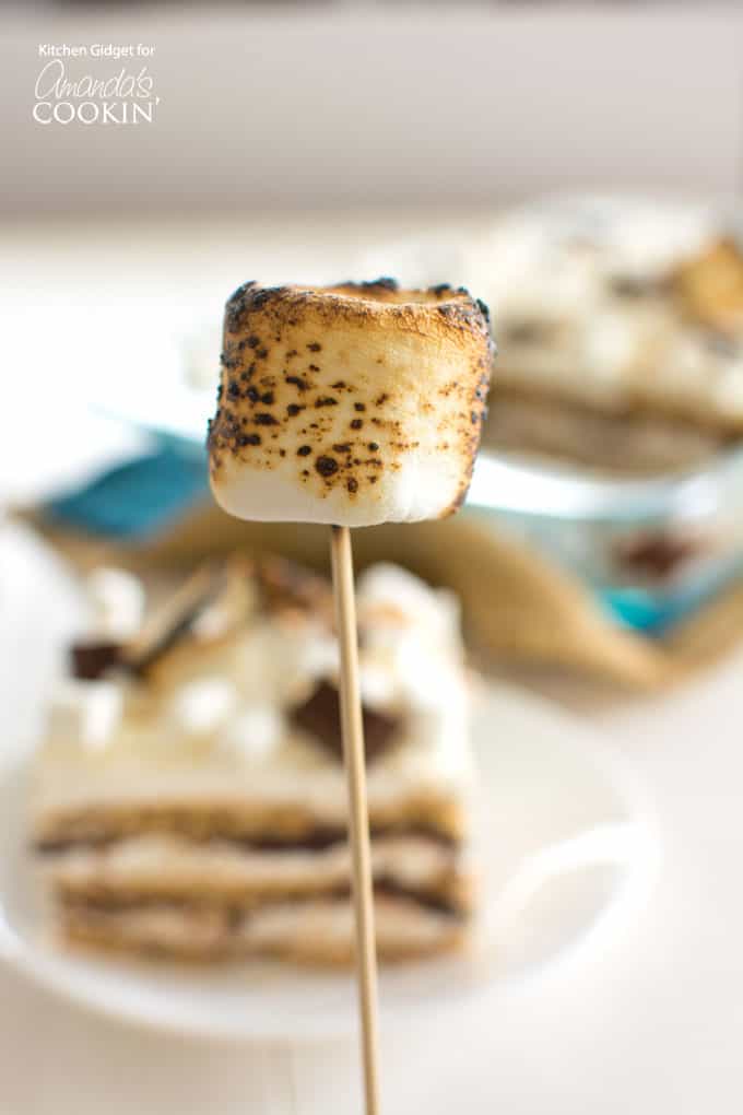 Toasted marshmallow on a stick in front of s'mores icebox cake