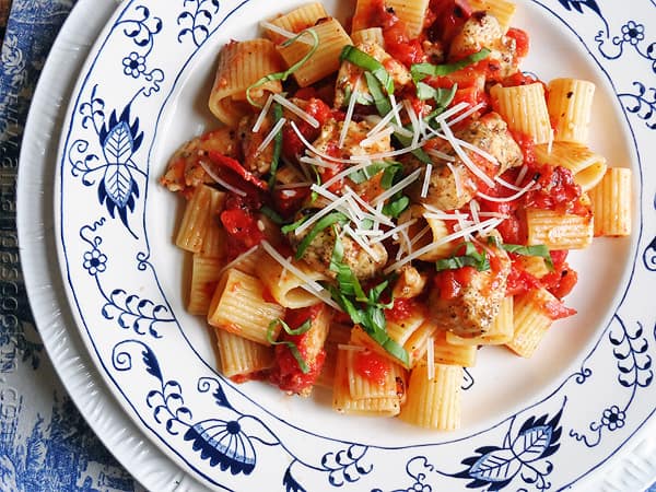 An overhead of a plate of spiced chicken and mezzi rigatoni topped with grated Parmigiano-Reggiano cheese and fresh basil.