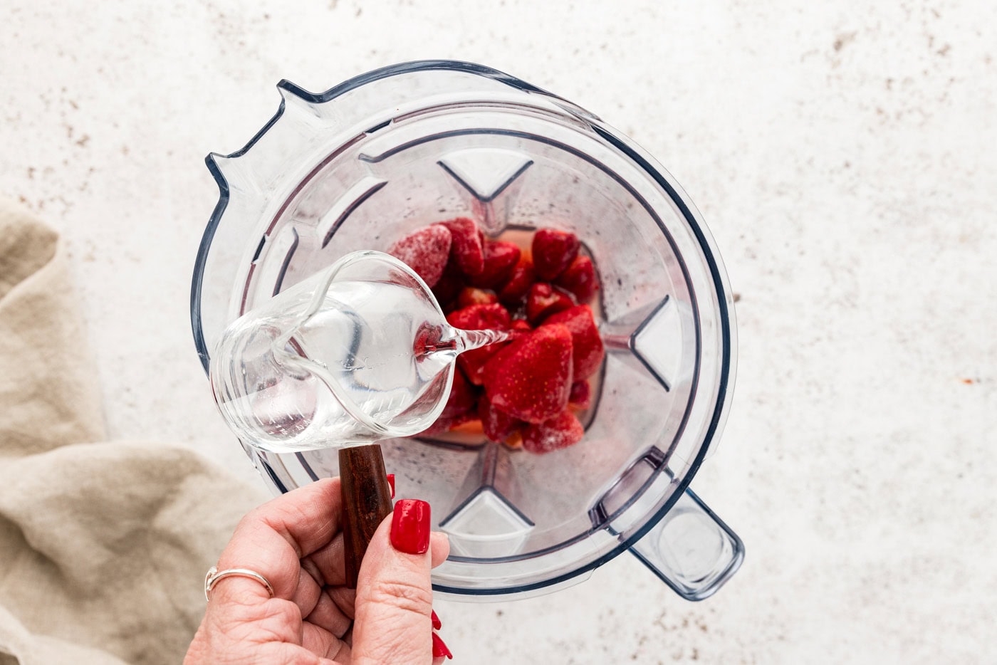 hand pouring silver tequila over top frozen strawberries in a blender