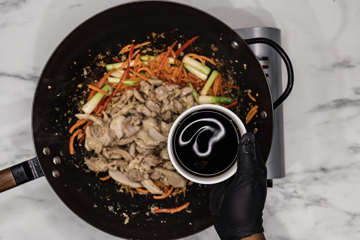 gloved hand holding a bowl of soy sauce over a skillet of chicken and stir fry veggies