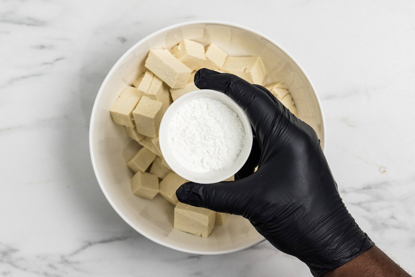 hand holding a bowl of cornstarch over cubed tofu in a bowl