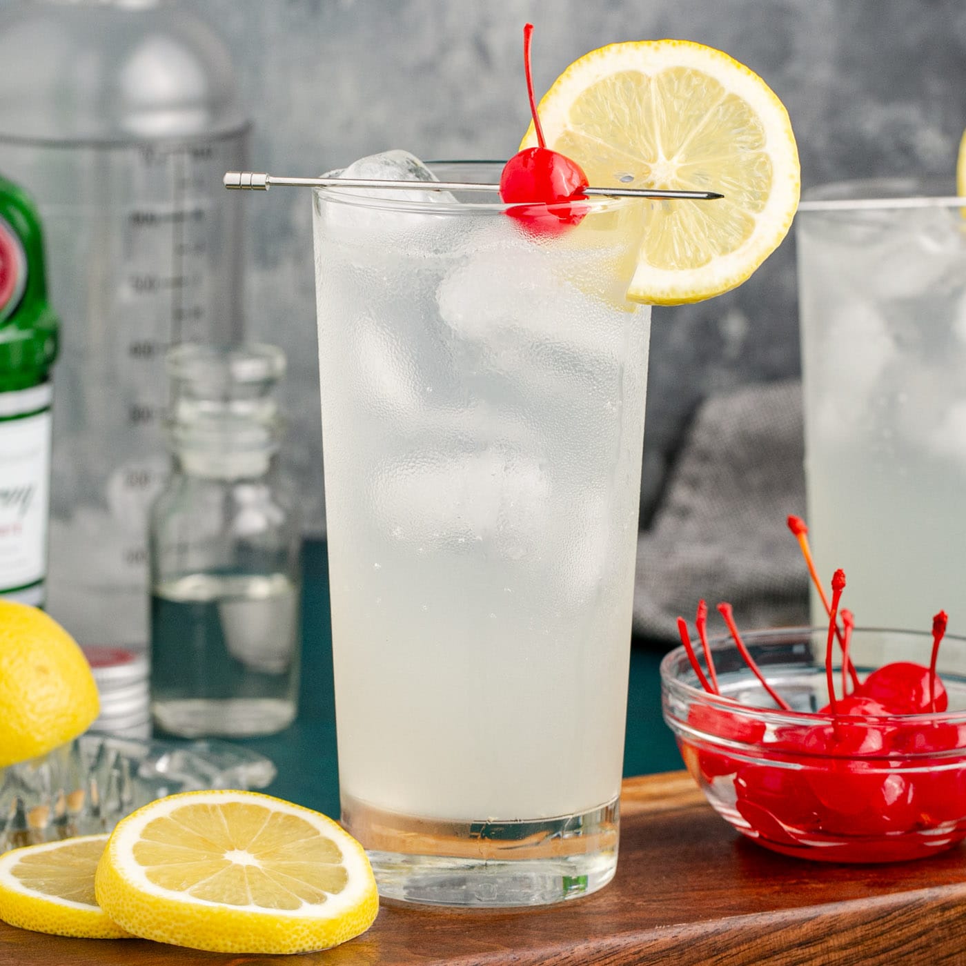 Tom Collins cocktail on a wooden board with lemon wheels and cherries on the side for garnish