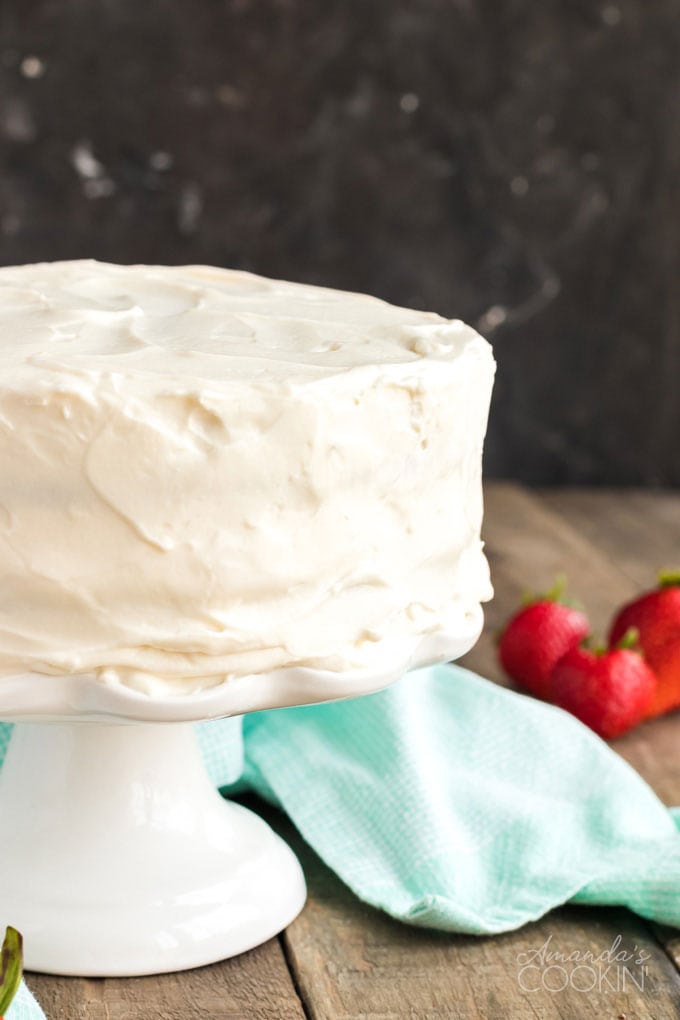 frosted white cake on a cake stand with a blue towel and strawberries in the background