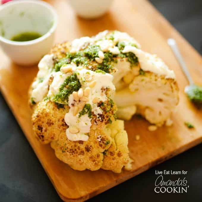 A close up of a whole roasted cauliflower topped with whipped feta and herb sauce on a wooden cutting board.