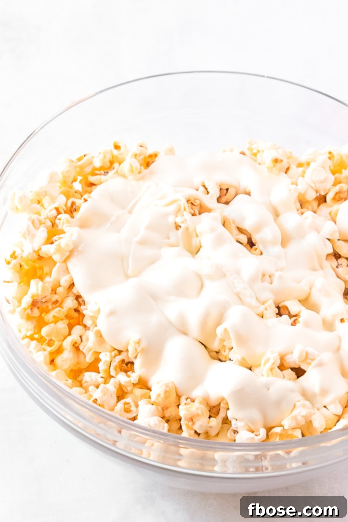 Melted white chocolate being poured over popcorn in a mixing bowl, ready to be stirred.