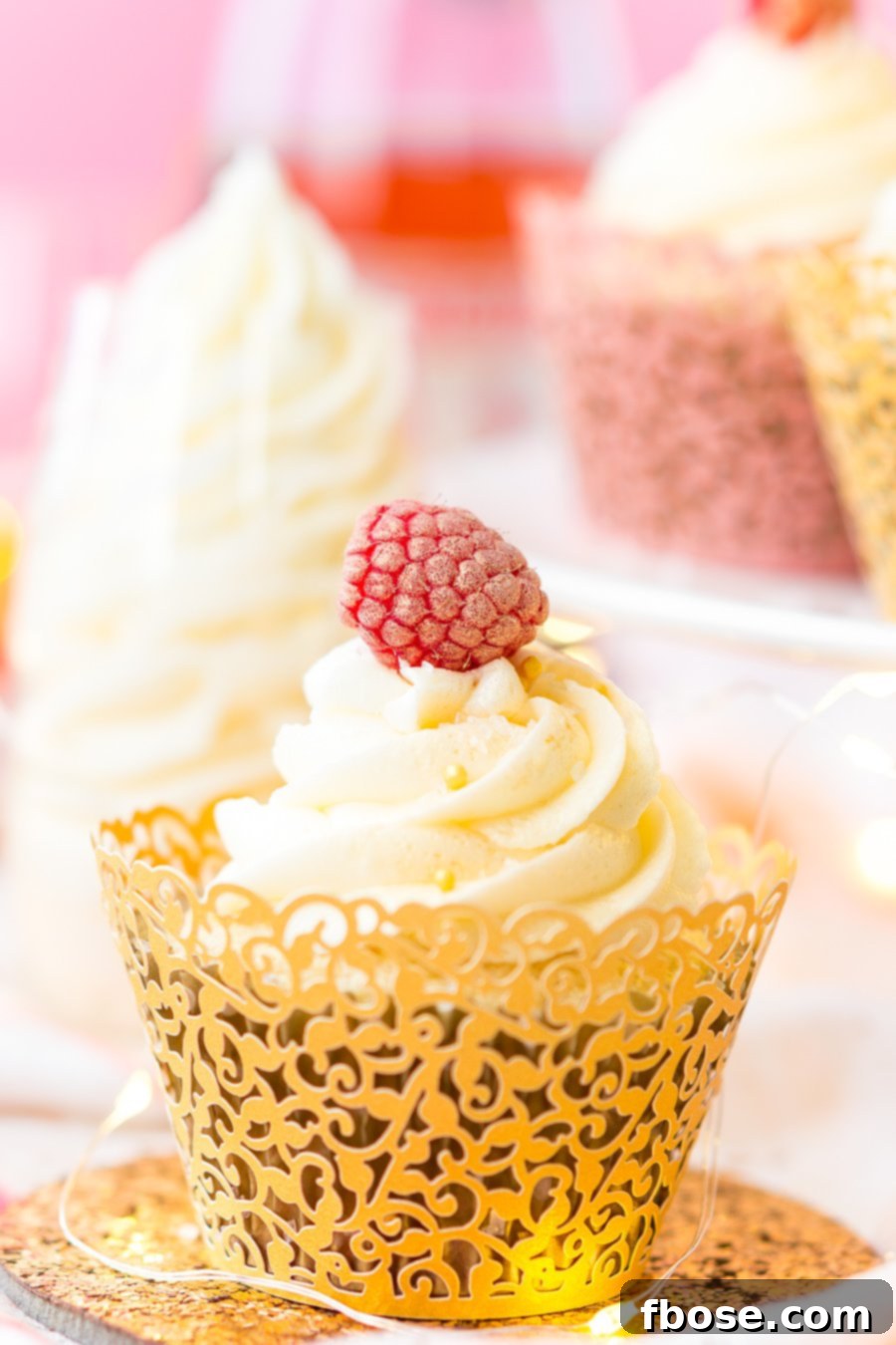 White Chocolate Raspberry Champagne Cupcakes, adorned with fresh raspberries and shimmering gold sprinkles, perfect for any festive occasion. The light cake and pink champagne buttercream promise a delightful treat.