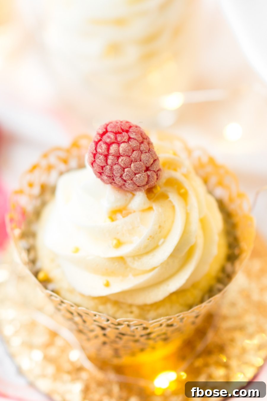 Three elegantly decorated White Chocolate Raspberry Champagne Cupcakes, ready for a party. Each cupcake features a fresh raspberry garnish and a dusting of gold shimmer on the buttercream.