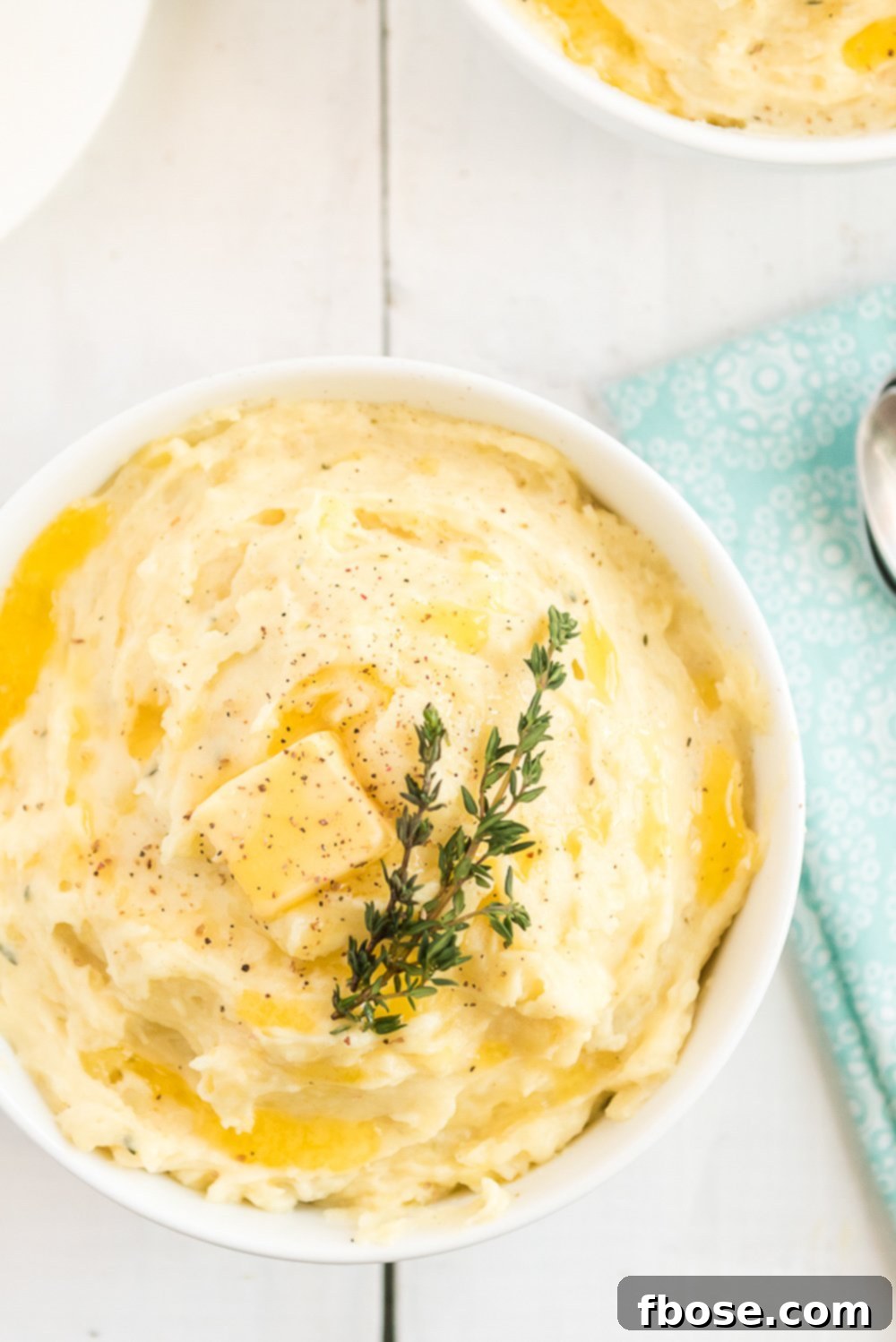 Overhead photo of Creamy Mashed Potatoes in a white bowl on white wood table with blue napkin.