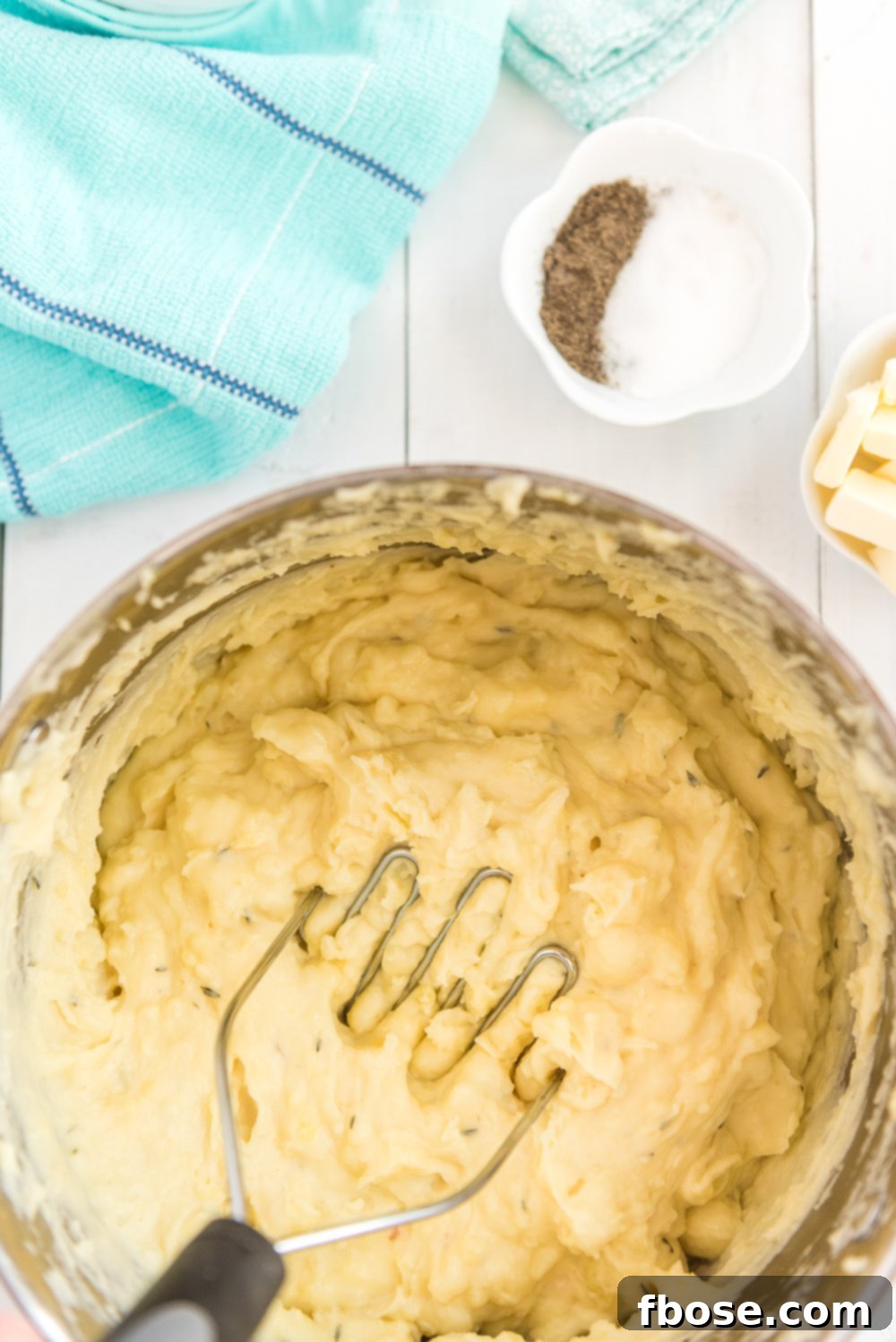 Overhead photo of boiled potatoes in large pot being mashed with a potato masher. Blue napkin and bowl of salt and pepper and butter to the side.