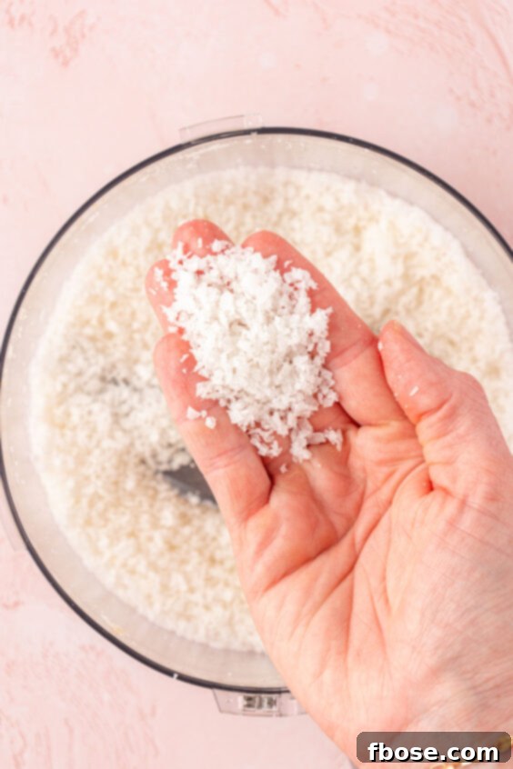 Coconut flakes in a food processor with a woman's hand holding some to the camera.