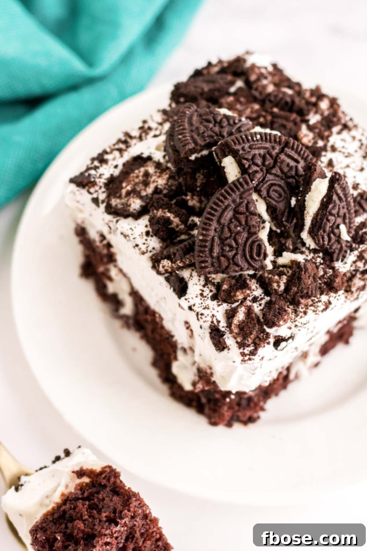 Slightly angled photo of a piece of Oreo chocolate cake on a white plate with a teal napkin next to it, showing the distinct layers.