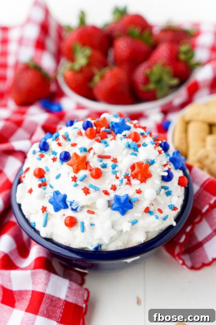 Red, White and Blue Cake Batter Dip 5 A close-up of a hand dipping a colorful sugar cookie into the Cake Batter Dip, highlighting the creamy texture and vibrant sprinkles.
