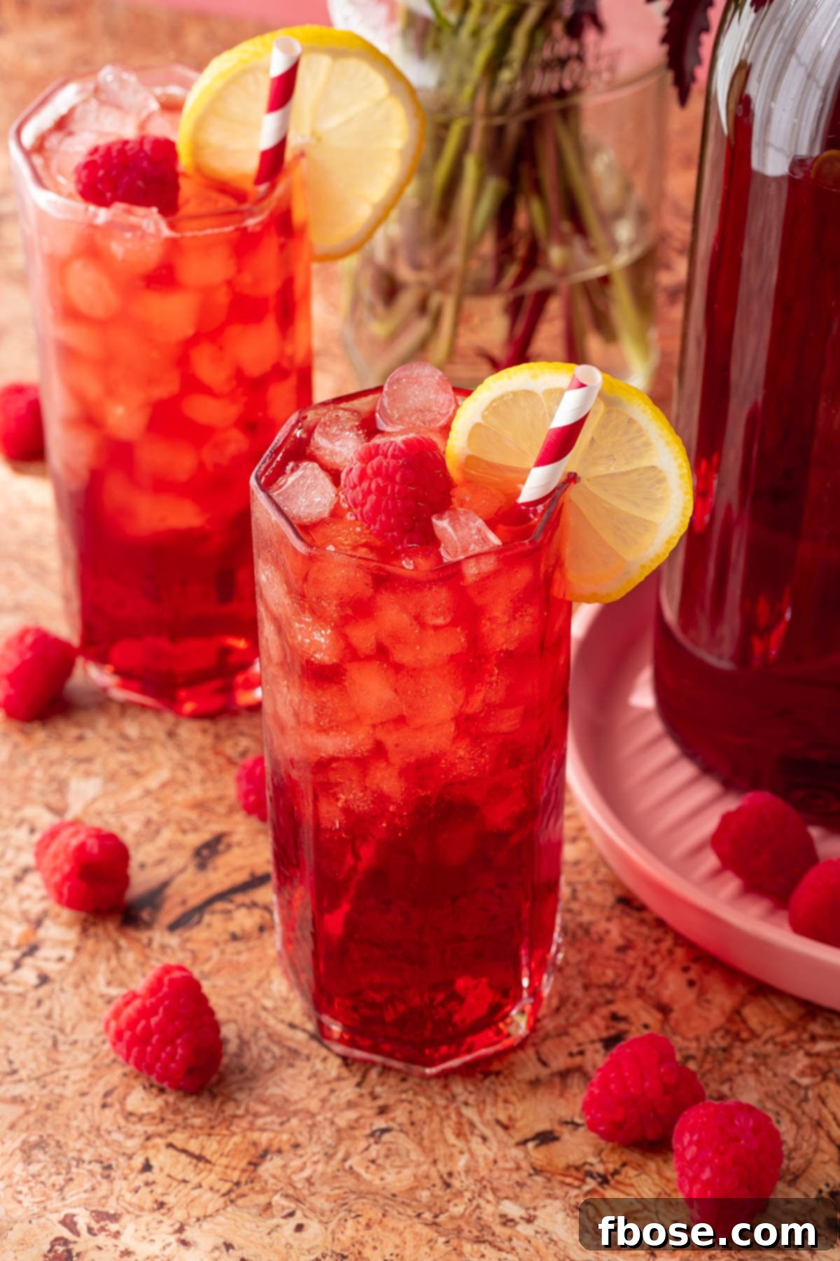 Two clear glasses of raspberry iced tea on a light-colored table, sparkling with ice and garnished with fresh raspberries and a sprig of mint.