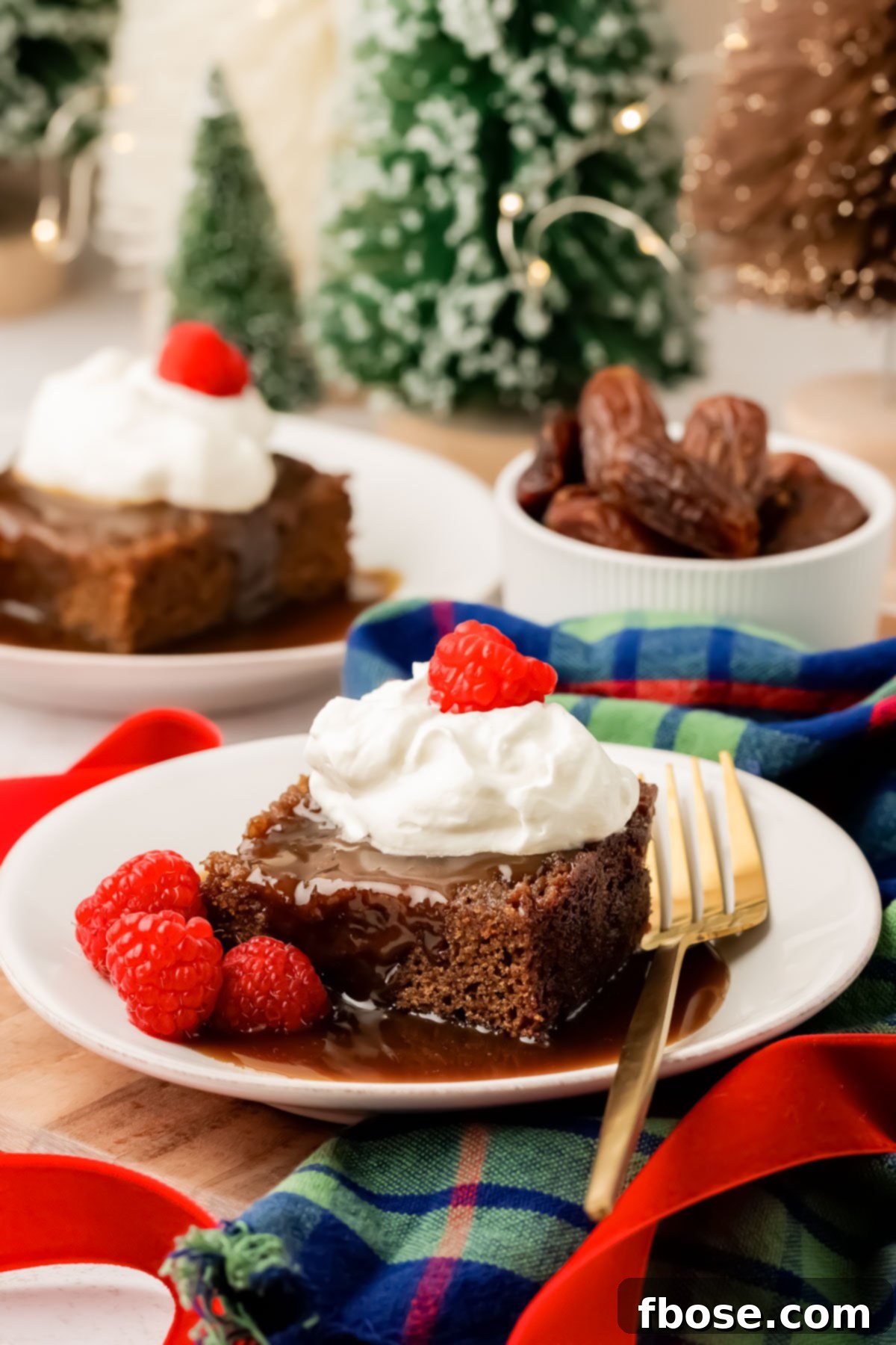 Slices of sticky toffee pudding on white plate with sauce, raspberries, and whipped cream.