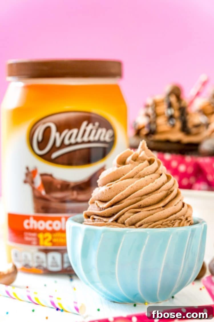 Small blue bowl filled with chocolate malt frosting with a container of Ovaltine and cupcakes in the background.
