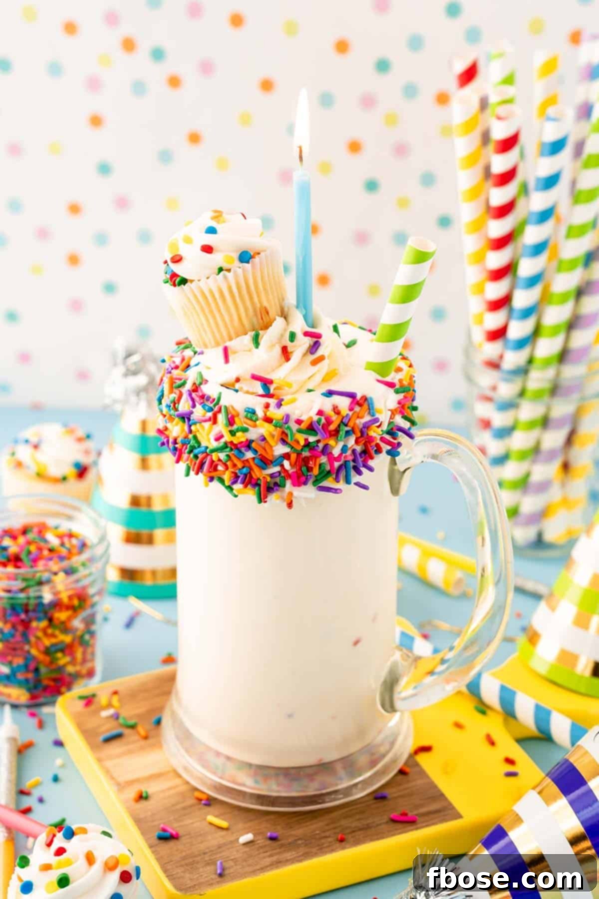 Close-up of a vibrant birthday cake milkshake in a tall glass mug, topped with whipped cream and sprinkles.