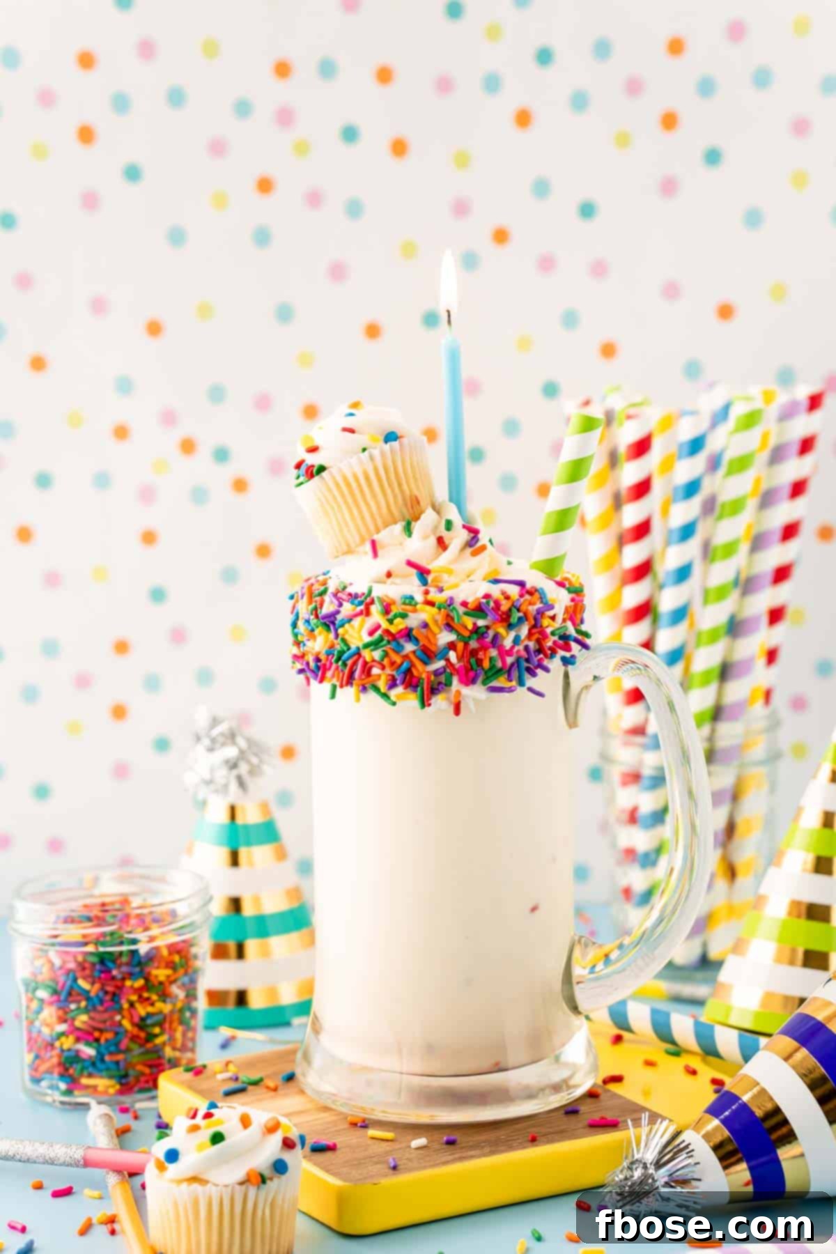 Another view of a birthday cake milkshake in a tall glass, garnished with a mini cupcake and sprinkles.