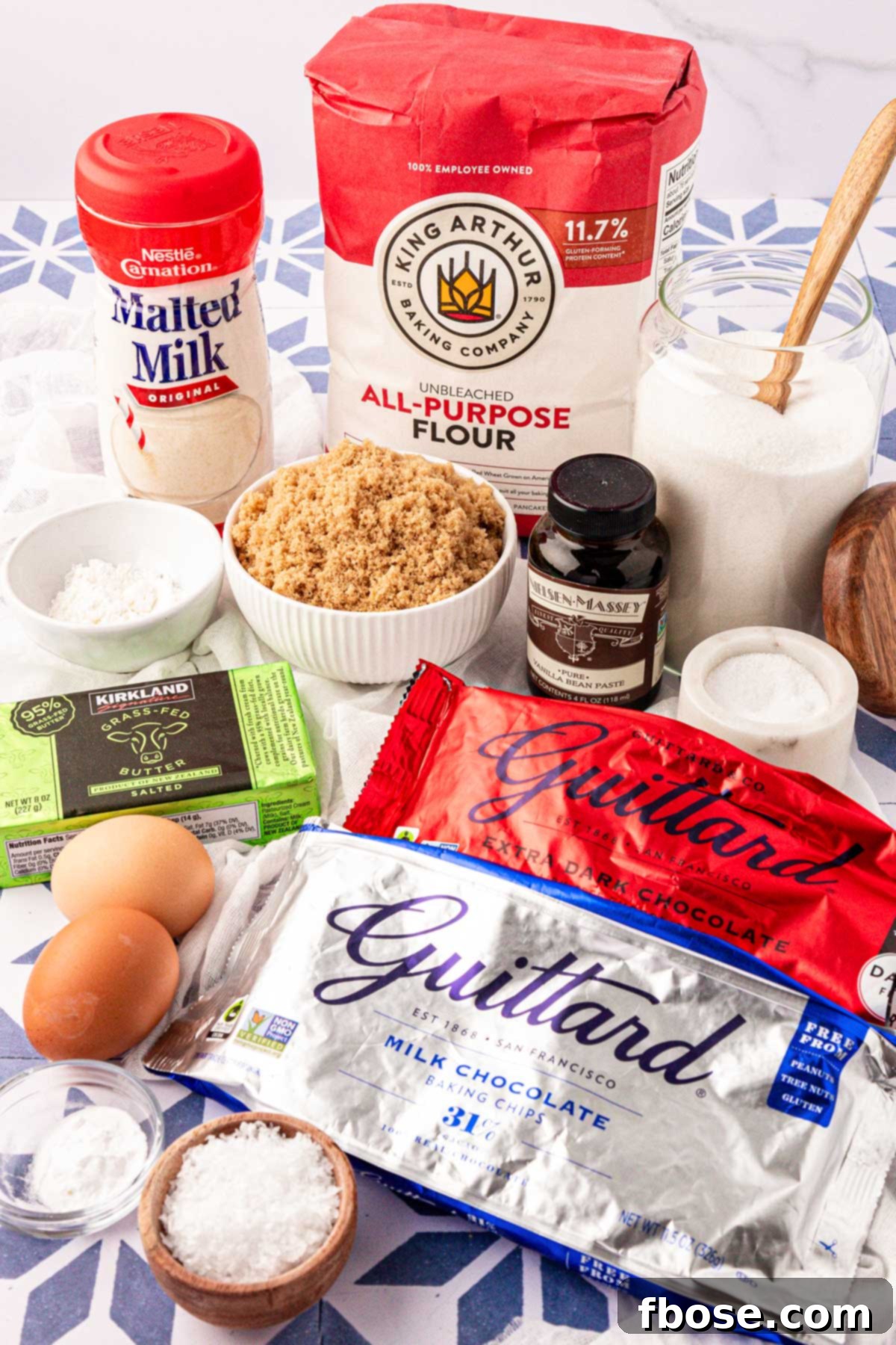 Assortment of chocolate chip cookie ingredients laid out on a table, including flour, sugars, butter, eggs, and various chocolate chips.