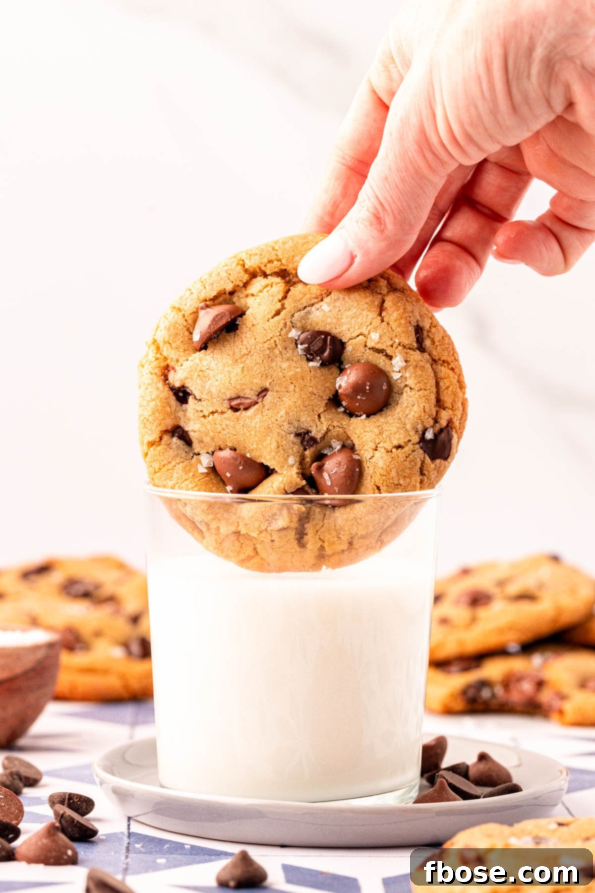 A woman's hand dipping a large, bakery-style chocolate chip cookie into a glass of cold milk, emphasizing its perfect texture.