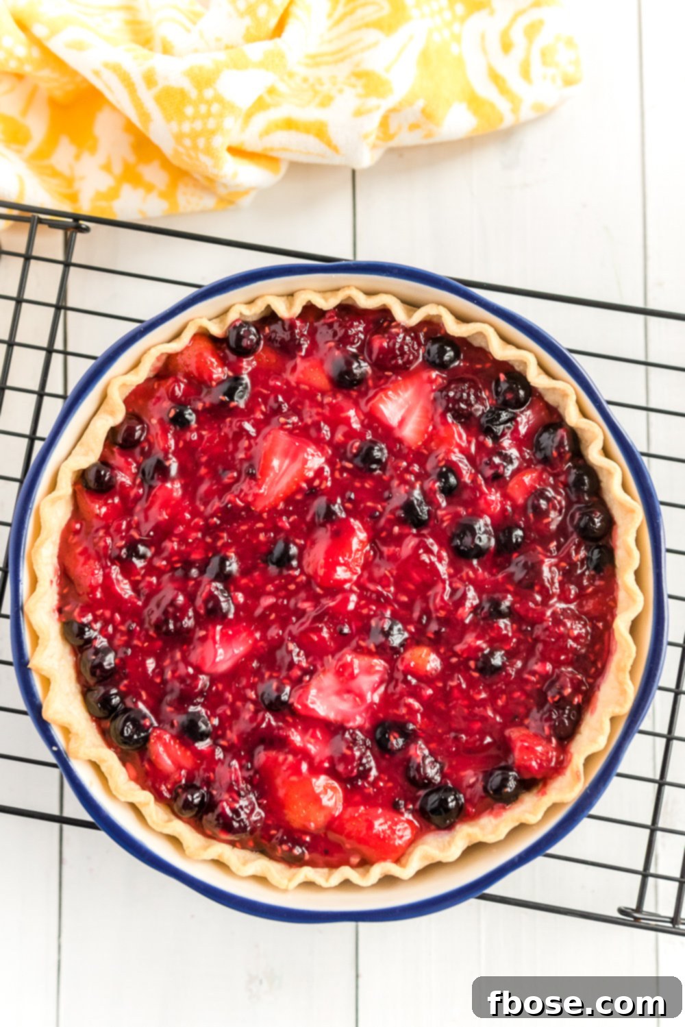 Mixed Berry Pie on black cooling rack on white wood with yellow napkin.