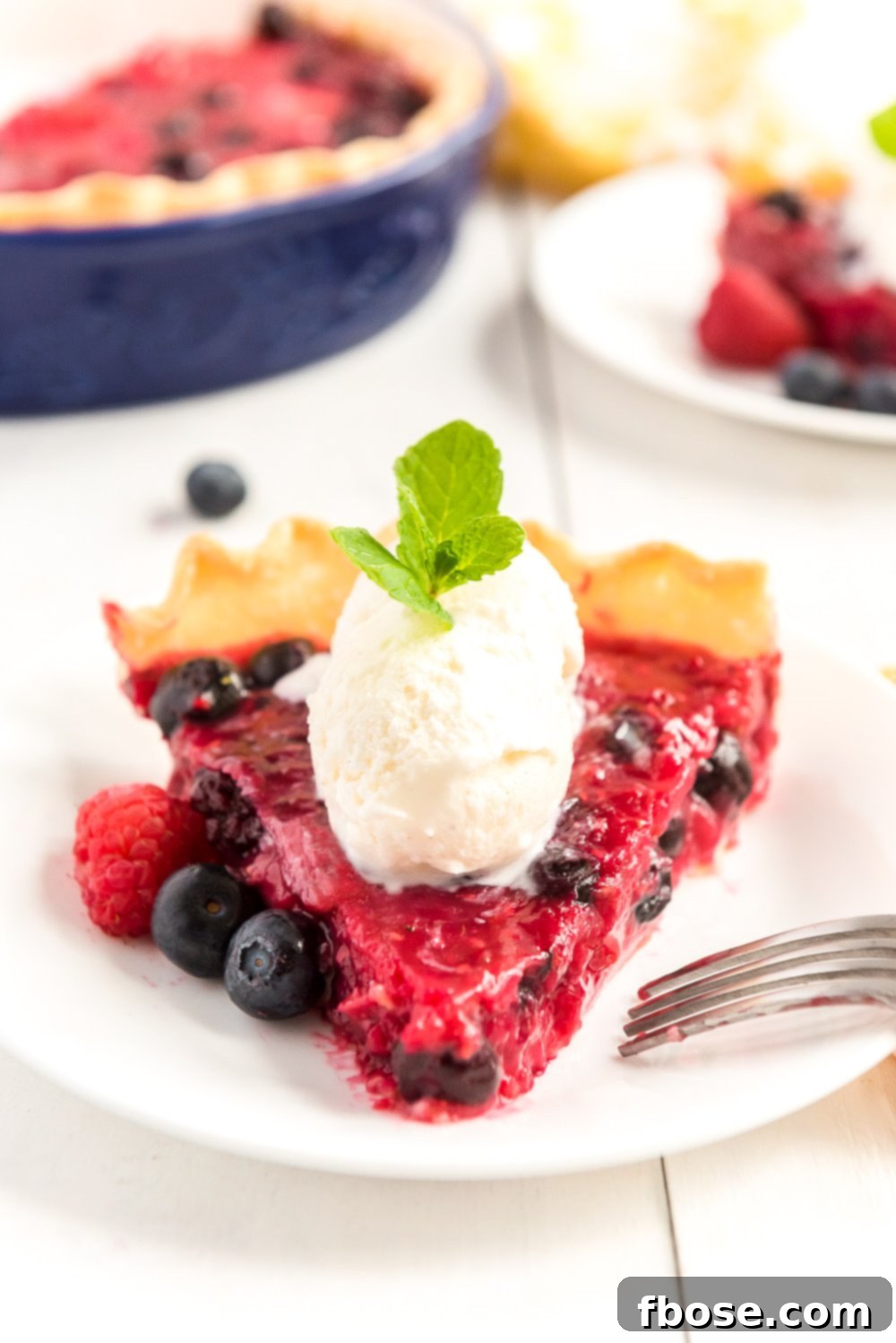 Straight on photo of a slice of Mixed Berry Pie pointed towards the camera with scoops of vanilla ice cream on top and fresh berries on the side.