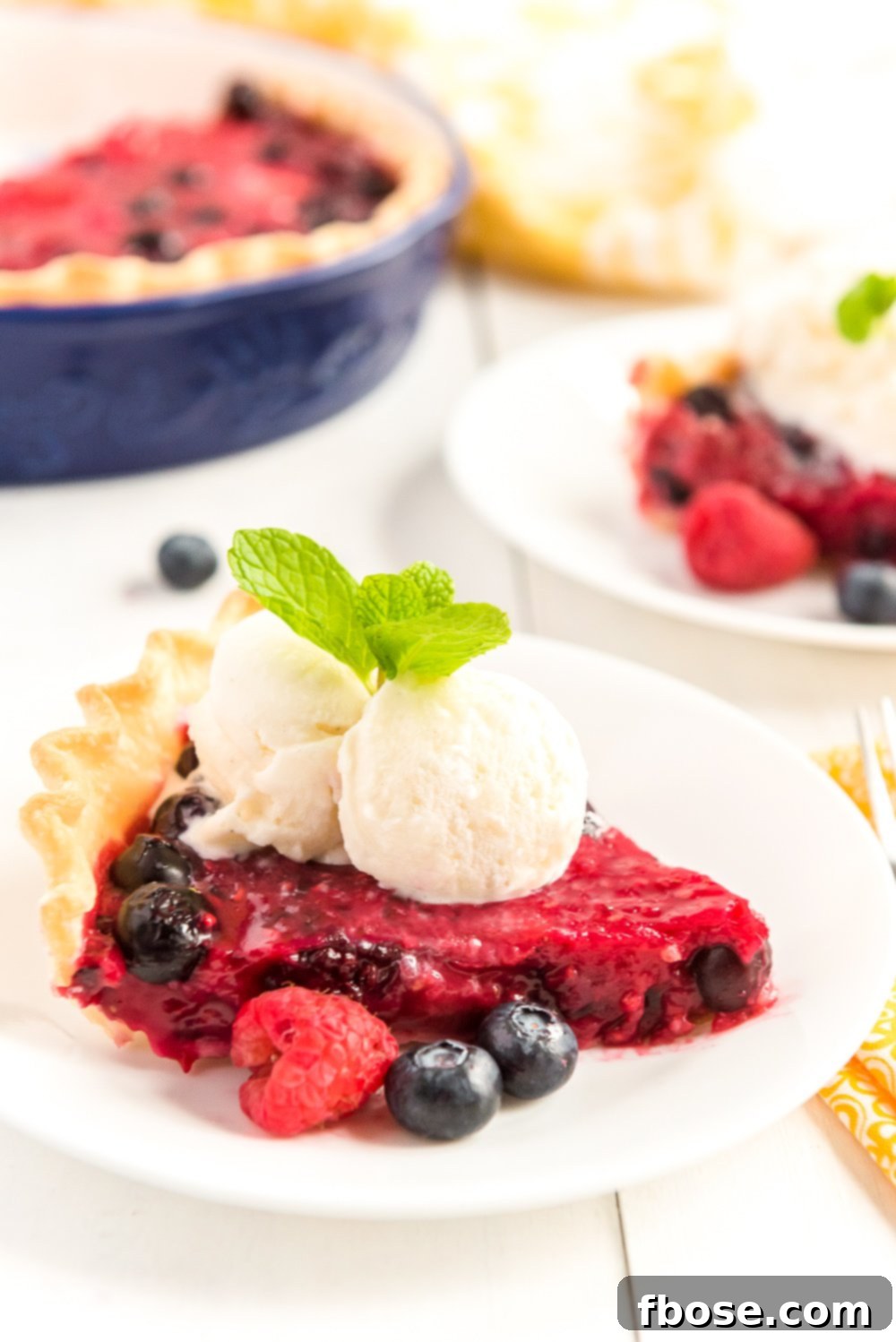 Slice of Mixed Berry Pie on a white plates with fresh berries, vanilla ice cream on top, and a sprig of mint.