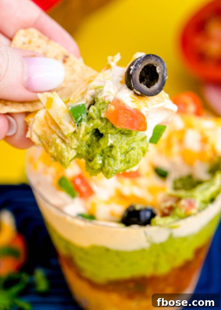 A woman's hand scooping a tortilla chip into a cup with 7 layer dip, showing the vibrant layers inside.