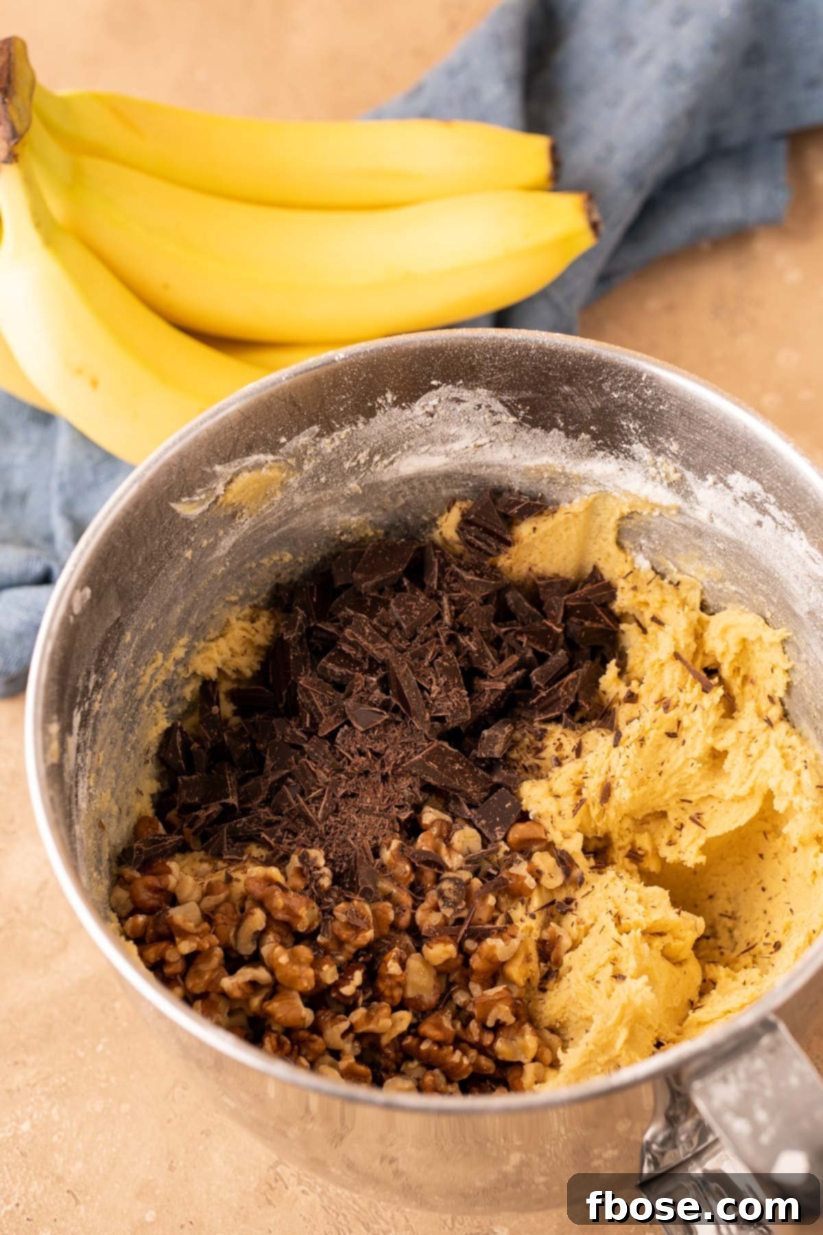 Decadent Chunky Monkey Pudding Cookies 9 Walnut and chopped chocolate being added to a stand mixer bowl filled with cookie dough.