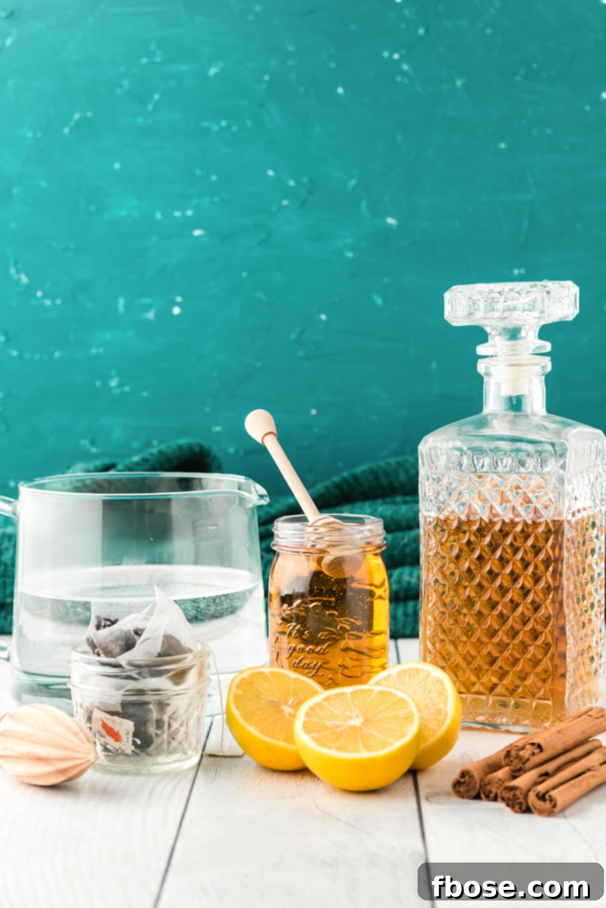 Ingredients to make a hot toddy laid out on a white wood table, including lemon, honey, whiskey, and tea.
