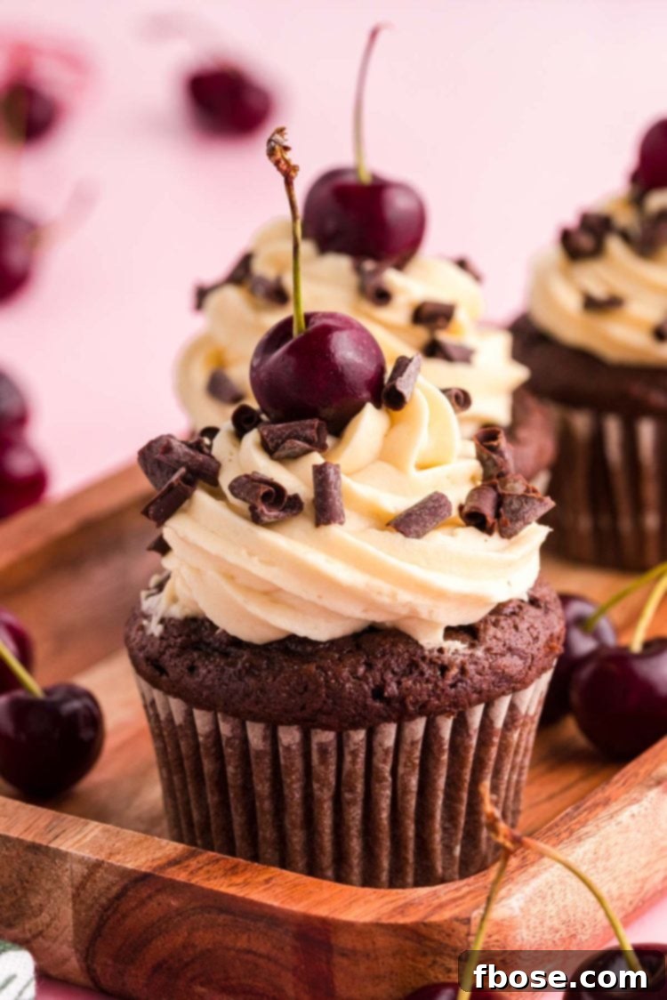 Close-up photo of a row of Black Forest Cupcakes adorned with fresh cherries and chocolate curls, resting on a wooden tray with a vibrant pink backdrop.