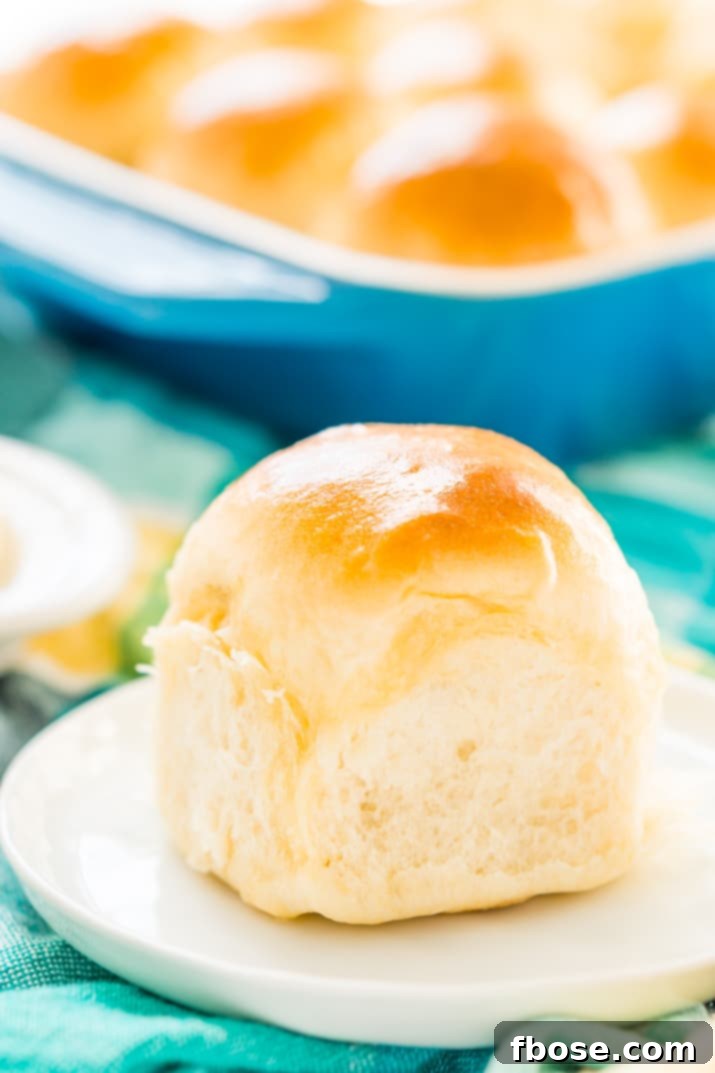 A single warm yeast roll placed on a small white plate, with a pan of more rolls blurred in the background.
