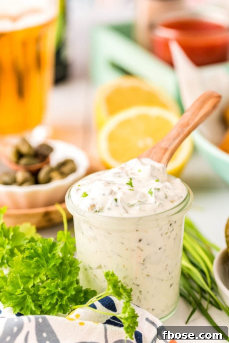 A close-up shot of a glass jar filled with vibrant homemade tartar sauce, a wooden spoon resting in it, with fresh pickles, lemons, and herbs blurred in the background.
