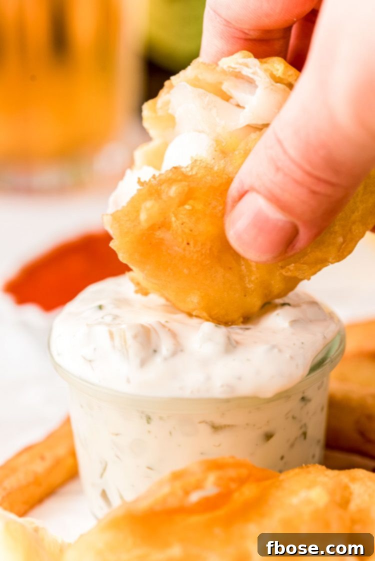 A person's hand gracefully dipping a piece of perfectly fried, golden-brown fish into a bowl of creamy, fresh homemade tartar sauce.
