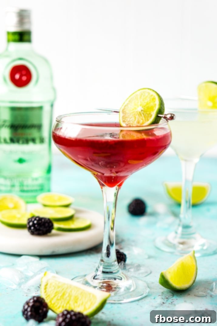 Blackberry Gimlet with a bottle of gin out of focus in the background, highlighting the cocktail's ingredients and inviting presentation.