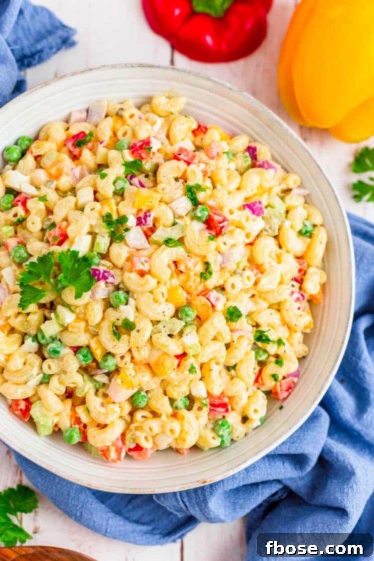 A large bowl of creamy classic macaroni salad, featuring tender elbow macaroni, colorful diced vegetables like red bell pepper, celery, and carrots, all coated in a rich, mayonnaise-based dressing. The salad is garnished with a sprinkle of paprika.